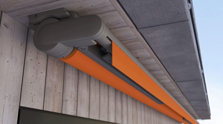 Retractable Awning Mx-930 With Orange Fabric Cover And Gray Brown Frame Is Attached To The Roof Overhang Of A Wooden House - Shade-Space