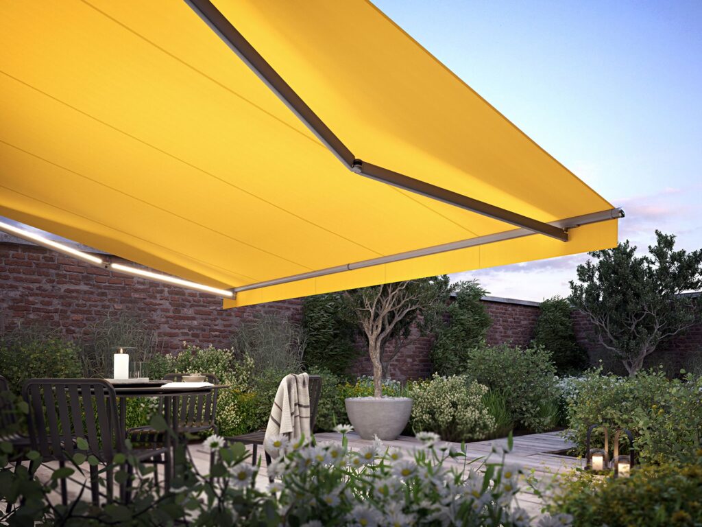 Yellow Fabric MX 1710 Awning Extended Over A Group Of Tables In A Small Garden The Led Lines On The Drop Arms Provide Light At Dusk - Shade-Space