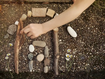 Child Arranging Pebbles And Sticks Creatively Into The Shape Of A House - Shade-Space